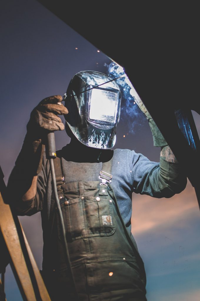 Worker wearing welding mask, gloves, and overalls holding metal sheet against a dusk sky, dark blue and orange tones