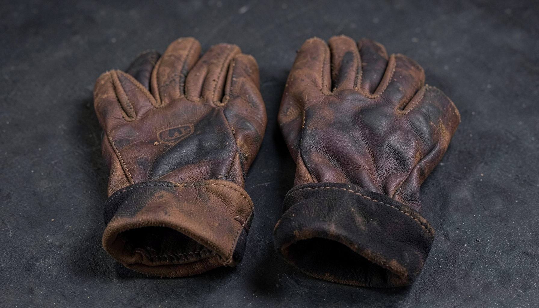 Pair of cowhide leather welding gloves with rolled cuffs on a dark surface, deep brown and charcoal tones, sturdy workshop feel