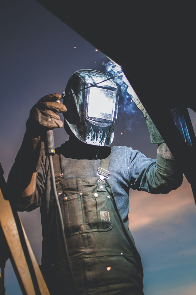 Worker wearing welding mask, gloves, and overalls holding metal sheet against a dusk sky, dark blue and orange tones
