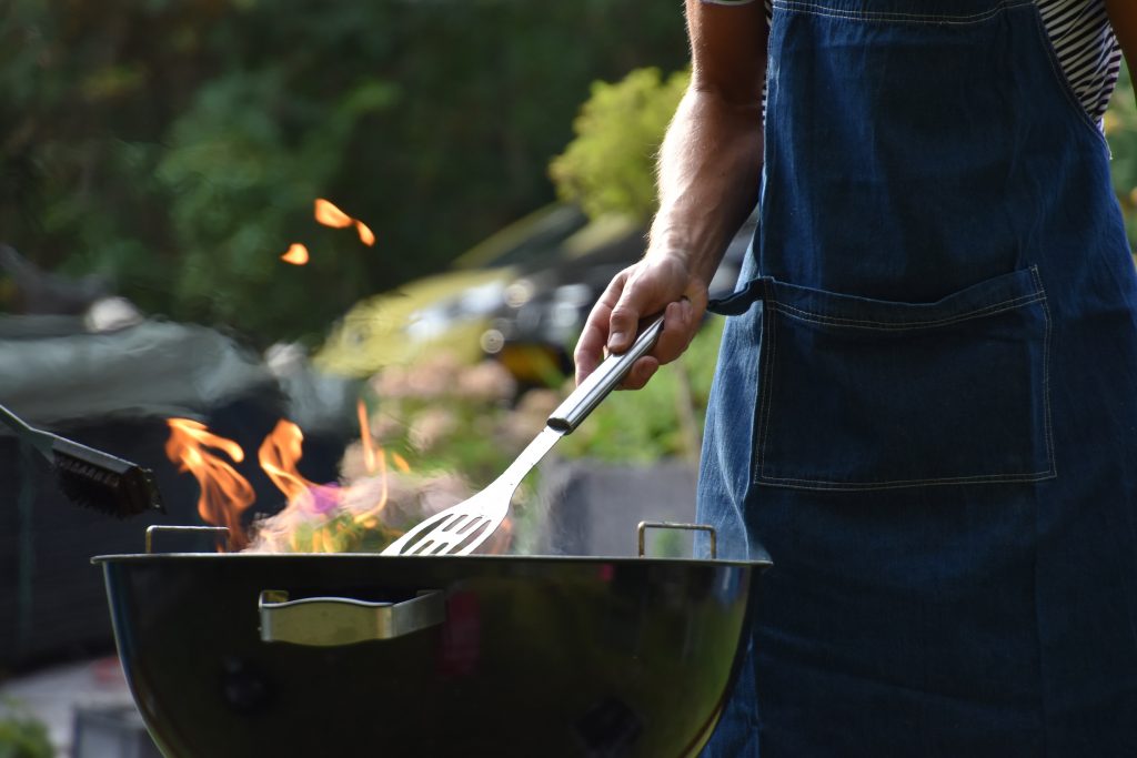 Person in denim apron grilling with flames rising from a barbecue, outdoor green background, casual mood