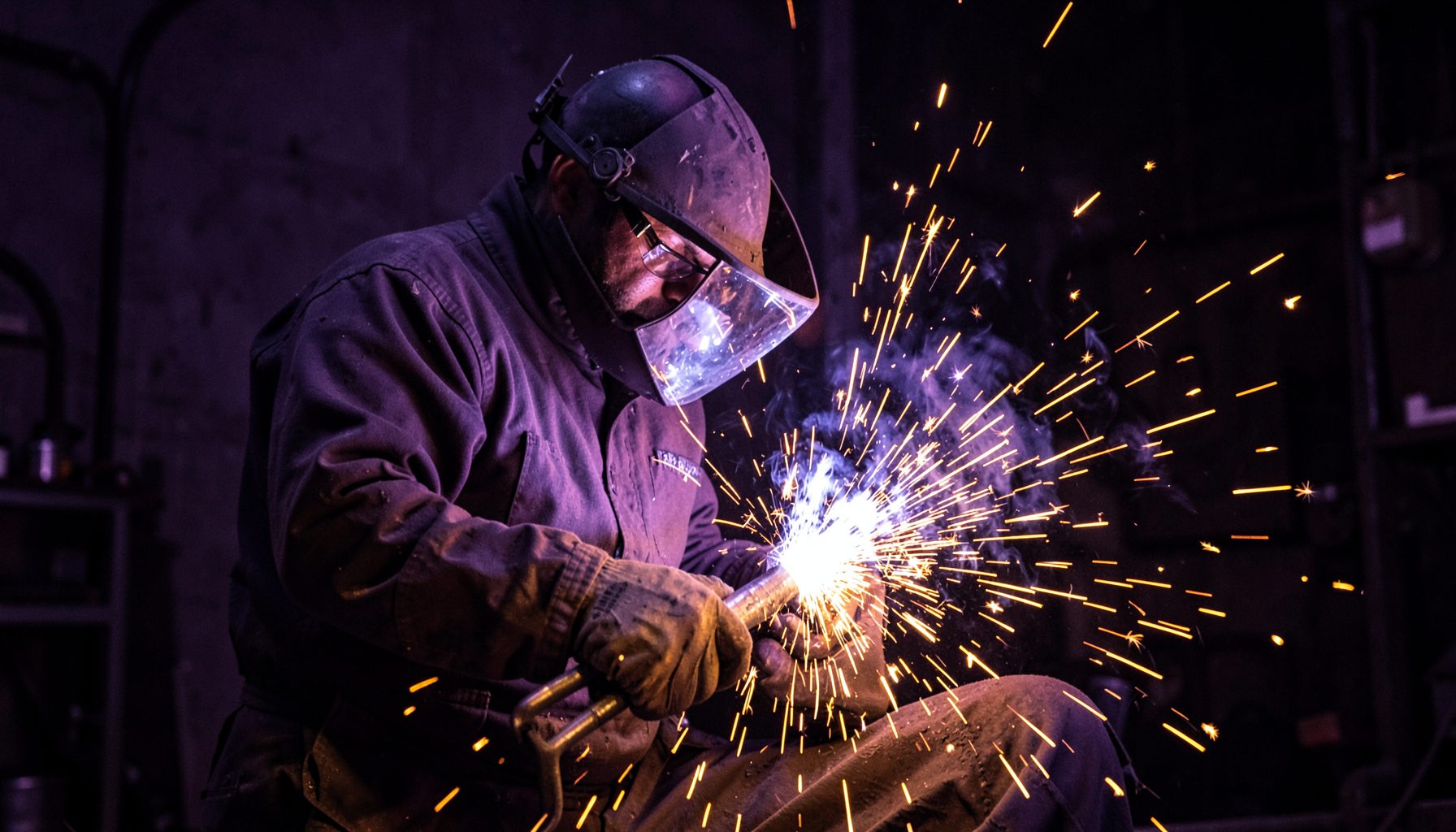 Welder in protective gear working with sparks flying against a dark industrial backdrop, deep purple and orange tones
